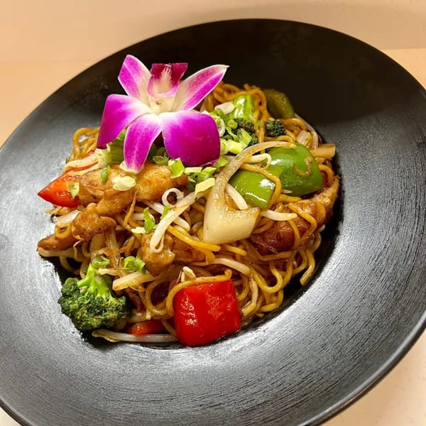 Japomen's ramen dish with noodles, chicken, colorful vegetables, and a purple orchid garnish in a black bowl.