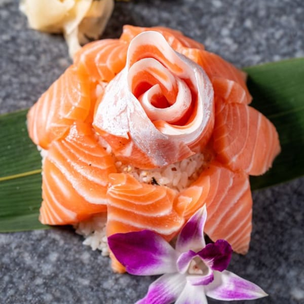 Salmon sashimi arranged like a rose on rice, garnished with a purple orchid and green leaf.