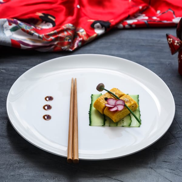 Tamagoyaki omelet rolls on cucumber slices, garnished with radish and chives, with chopsticks.